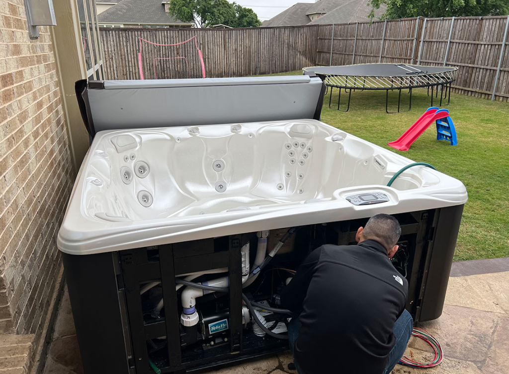 Back view of a technician servicing an open-panel hot tub in a backyard setting, showing the accessible electrical and plumbing components, and perimeter insulation.
